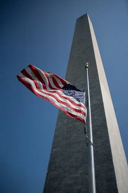 Low angle shot of the Washington Monument with the US flag waving beneath a clear blue sky.